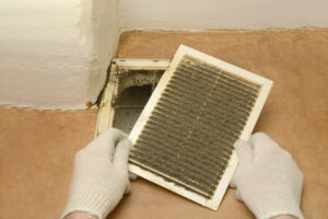 Pair of hands wearing white gloves, removing dust from dirty air duct vent in house.