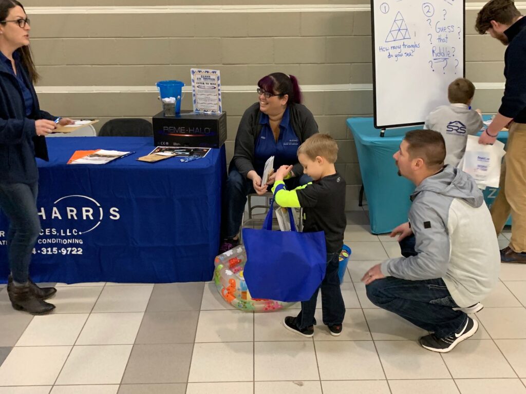 Harris Air staff at Harris Air table, giving free merchandise to small children holding tote bags.